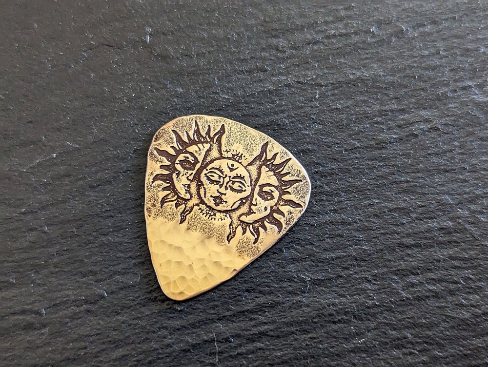Bronze guitar pick with suns and faces and a sparkling hammered texture -